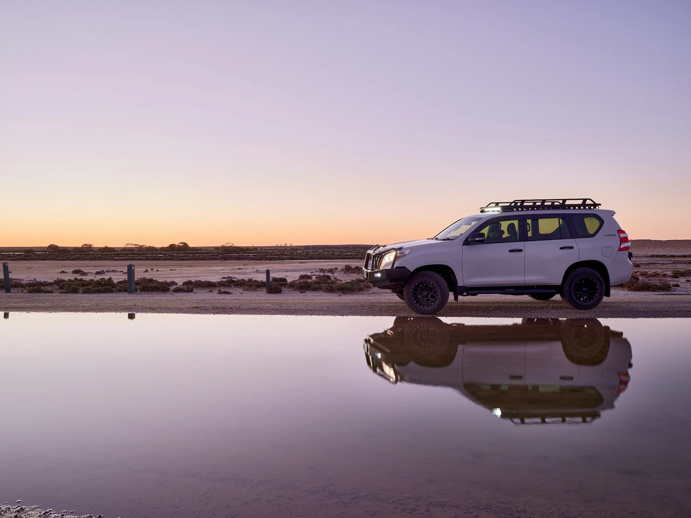 A white SUV with its lights on beside still water at dusk